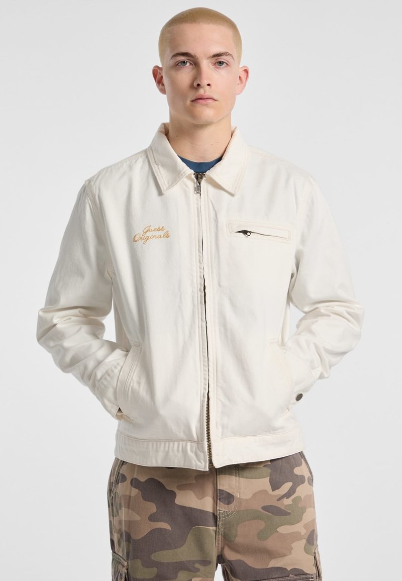 White canvas jacket with a zip closure, stand collar, and left chest pocket. Features "Guess Originals" in gold embroidery.