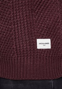 Primo piano di un maglione lavorato a maglia color marsala con un'etichetta bianca rettangolare "JACK & JONES CORE" cucita sul lato anteriore in basso sopra dei pantaloni neri.