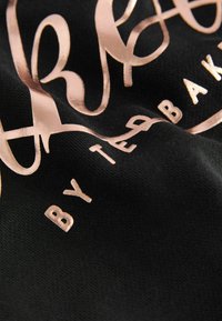Černá textilie s kovovým růžovým zlatým písmem, které zní "Ted Baker", a plynulou typografií, zdůrazňující texturu a kontrast.