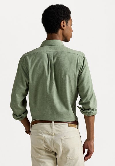 Chemise verte à boutons avec manches retroussées, tissu texturé et pli dans le dos, associée à un pantalon de couleur claire et une ceinture marron.