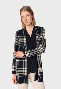 Navy och krämrandig cardigan med avslappnad passform, långa ärmar, öppen fram och texturerad tyg. Bärs över en mörk topp och krämfärgade byxor.