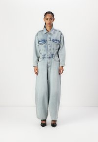 Denim jumpsuit i lys blå med en strukturert, belted midje, vide ben og to brystlommer; brukt med svarte spisse hæler.