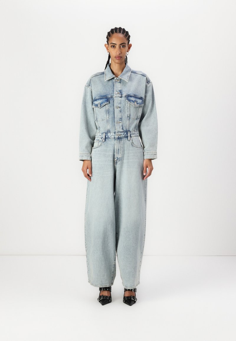 Denim jumpsuit i lys blå med en strukturert, belted midje, vide ben og to brystlommer; brukt med svarte spisse hæler.