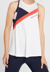 Camiseta sin mangas deportiva en blanco, con rayas diagonales en azul marino y coral brillante. Cuenta con un panel de malla transpirable en el escote.