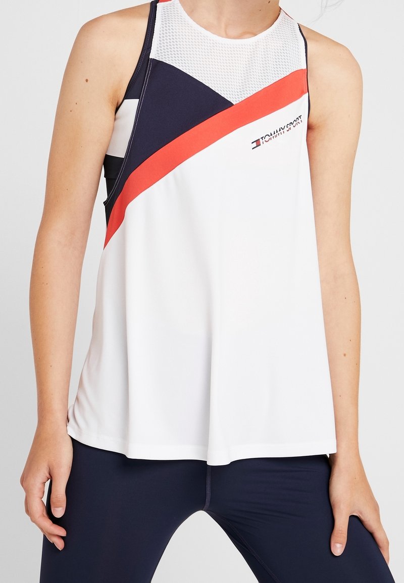 Camiseta sin mangas deportiva en blanco, con rayas diagonales en azul marino y coral brillante. Cuenta con un panel de malla transpirable en el escote.