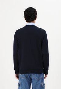 Sweatshirt blu navy con maniche lunghe e scollatura rotonda, realizzato in tessuto liscio, presenta un orlo e polsini a coste, indossato sopra una camicia azzurro chiaro.