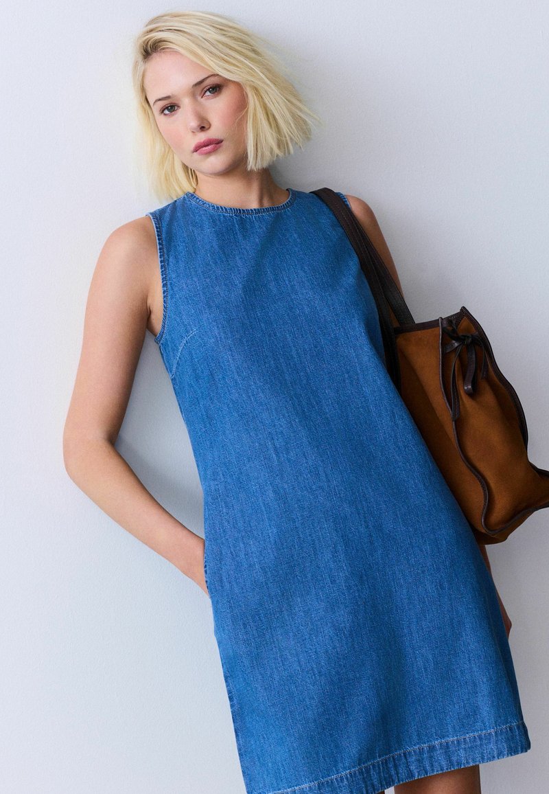 Jeune femme aux cheveux blonds courts porte une robe en jean bleue sans manches, avec un sac à bandoulière marron, debout contre un mur gris clair.