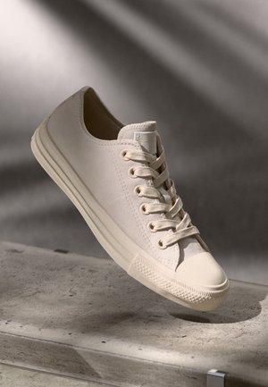 Beige canvas sneaker met veters en rubberen zool, rustend op een betonnen oppervlak onder zacht licht.
