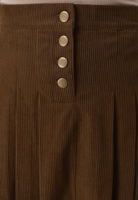 Καφέ φούστα από ριμπ ύφασμα με τέσσερα στρογγυλά ορειχάλκινα κουμπιά τοποθετημένα κάθετα κοντά στη μέση και μία ελαφριά πτυχή στη δεξιά πλευρά.