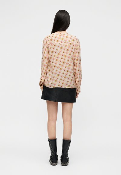 Γυναίκα με μακριά σκούρα μαλλιά, ντυμένη με ροζ φλοράλ μπλούζα με μακριά μανίκια, μαύρη μίνι φούστα και μαύρες μπότες μέχρι τη μέση της γάμπας, που στέκεται με την πλάτη προς τα εμπρός.