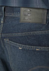 Denimjeans i mörkblått med en texturerad läderlapp märkt "G-STAR RAW." Har bakfickor och kontrasterande sömnadsdetaljer.