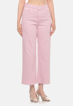 Donna che indossa pantaloni a vita alta, a gamba larga, rosa chiaro, con scarpe beige con tacco alto e dettagli ritagliati, in piedi su sfondo bianco.