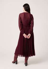 Burgunderfarbenes, plissiertes Maxikleid mit transparenten Ärmeln, elastischem Bund und dezentem Glanz. Kombiniert mit knielangen burgunderfarbenen Absatzstiefeln.