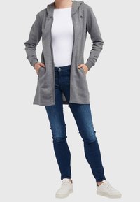Grijze rits hoodie met een capuchon en voorkeurszakken, gecombineerd met blauwe skinny jeans en witte sneakers. Eenvoudig ontwerp, zachte textuur.