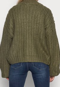 Eine Person trägt einen übergroßen olivgrünen, grob gestrickten Pullover und dunkelblaue, figurbetonte Jeans, von hinten vor einem einfarbigen Hintergrund gezeigt.