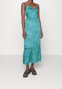 Robe sans manches, couleur teal, avec un motif à rayures zèbre, un détail en trou de serrure au niveau du col et un ourlet froncé. Portée avec des sandales noires à semelles épaisses.