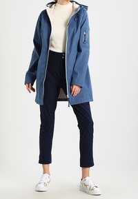 Blå lång jacka med huva, ficka med dragkedja, lätt foder, bärs över en krämfärgad tröja och marinblå byxor. Vita sneakers med blommiga detaljer.