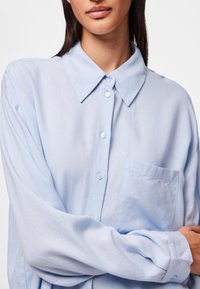 Camisa de botones azul claro hecha de una tela suave, con cuello, mangas largas y un bolsillo en el pecho con una sutil textura.