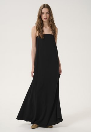 Robe maxi noire avec encolure droite, tissu léger, design fluide et fentes latérales. Femme portant des chaussures beiges se tient devant un fond blanc.