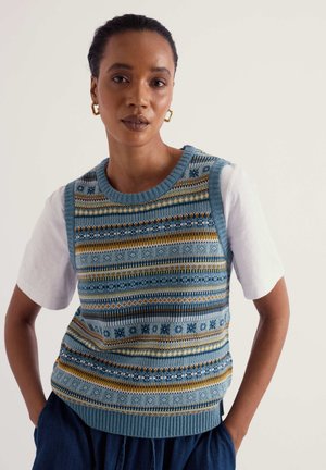Mujer con el pelo recogido lleva una camiseta blanca, un chaleco de punto azul con estampado, pantalones oscuros y pendientes de aro dorados, de pie sobre un fondo liso.
