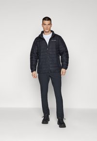 Giubbotto a vento nero con zip frontale e colletto rialzato, caratterizzato da motivi a righe orizzontali, indossato sopra una camicia bianca e abbinato a pantaloni scuri.
