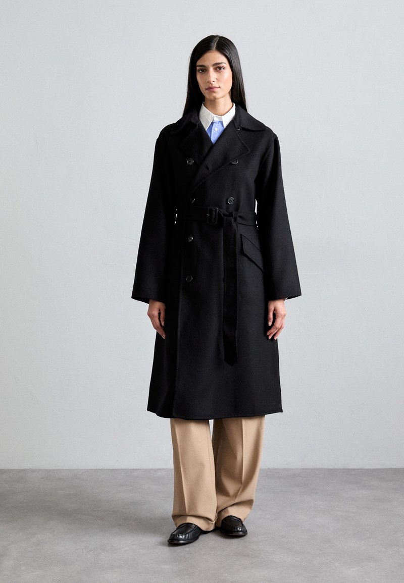 Μαύρο παλτό από μάλλινο ύφασμα με ντούμπλα, φαρδιές πέτες, ζώνη και μπροστινές τσέπες, φορεμένο πάνω από μπεζ παντελόνι φάρδους.