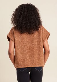 Brauner, strukturierter Pullover mit kurzen Ärmeln und lockerer Passform. Enthält kleine Farbtupfer in helleren Nuancen und wird über dunklen Jeans getragen.