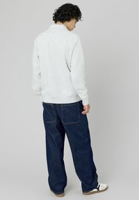 Personne avec des cheveux bouclés portant un sweat-shirt gris clair, un jean bleu foncé ample, et des baskets blanches, se tenant de dos contre un fond uniforme.