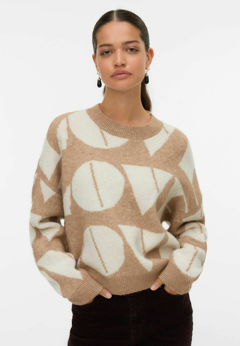 Pull en beige et crème avec des motifs géométriques, fabriqué en tissu doux, doté d'un col rond et de poignets côtelés, associé à un pantalon noir.