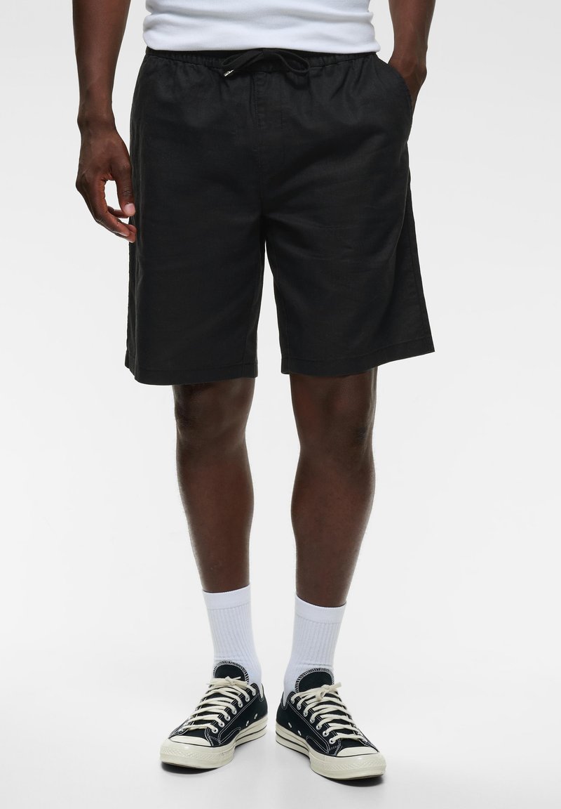 Homme portant un short noir à cordon, des chaussettes blanches, des baskets noires basses, debout les mains dans les poches sur fond blanc.