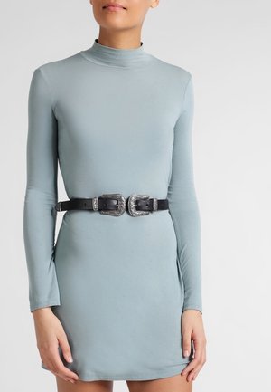 Femme portant une robe ajustée à manches longues bleu clair avec une ceinture noire à double boucle argentée à la taille, les mains reposant le long du corps.