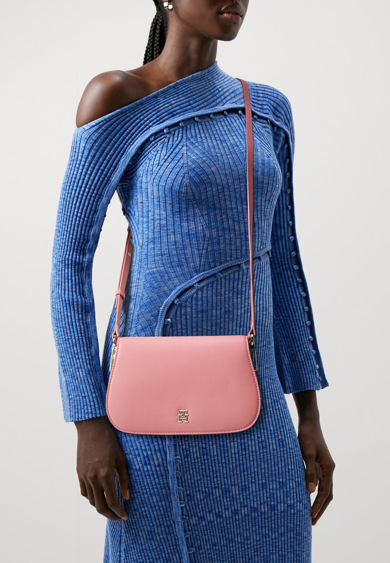 Rosa Leder-Umhängetasche mit glatter Textur, ausgestattet mit einem goldfarbenen Logo, einem schmalen Trageriemen und rechteckiger Form. Trägt man über einem blauen gerippten Kleid.