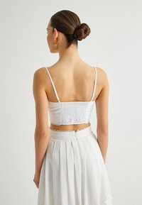 Witte crop top met verstelbare dunne bandjes en smocktextuur aan de achterkant, gecombineerd met een witte geplooide rok met decoratieve accenten.