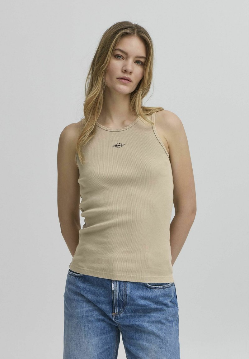 Top de tirantes de canalé beige con cuello redondo, tirantes finos y un pequeño logo negro bordado en el pecho. Combinado con unos jeans de mezclilla azules.