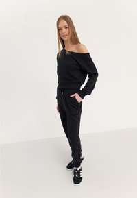 Sweat-shirt noir décontracté à épaules dénudées, associé à des joggers noirs, avec une taille ajustable et des poches latérales. Le modèle porte des baskets noires.