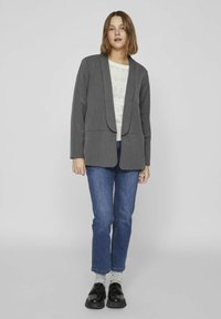 Blazer gris ajusté avec un col à revers, deux poches avant et un design ouvert, porté par-dessus un haut à motifs clair et un jean bleu.