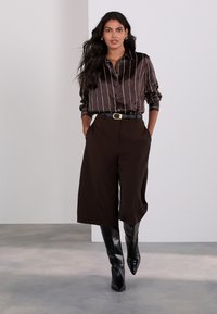 Braunes Satin-Hemd mit Knöpfen und weißen Streifen, kombiniert mit brauner Hose mit weitem Bein und glänzenden schwarzen kniehohen Stiefeln. Detail mit goldener Schnalle am Gürtel.