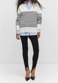 Femme portant un pull blanc à rayures bleu marine, une chemise bleu clair, un pantalon noir ajusté et des escarpins noirs pointus avec des brides à la cheville, debout.