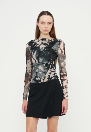 Femme aux longs cheveux bruns portant un haut à manches longues noir et blanc à motifs et une jupe portefeuille noire, debout devant un fond uni.