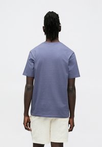 Maglietta di cotone viola con maniche corte, collo rotondo e trama liscia, indossata con pantaloncini bianchi con motivo. Vista posteriore.