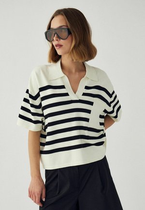 Naine, kes kannab üle suurusega mustasid päikeseprille, lühikese varrukaga valget polo särki mustade horisontaalsete triipudega ja musti pükse, seisab ühevärvilise tausta ees.