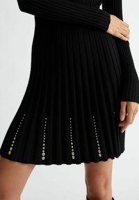 Robe noire à côtes avec manches longues, présentant des plis verticaux et des embellissements de paillettes rondes le long de l'ourlet.