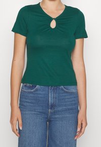 Vihreä lyhythihainen t-paita, jossa avainreikämuotoilu pääntiessä ja kerätty tekstuuri. Yhdistetty sinisiin korkeavyötäröisiin farkkuihin.