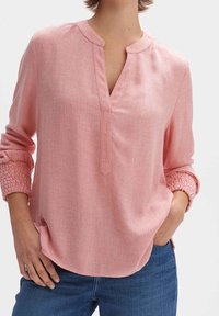 Blouse rose clair avec un col en V et une patte de boutonnage, dotée de manches froncées et d'un tissu doux et texturé. Idéale pour une tenue décontractée.