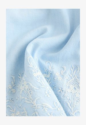 Tissu bleu clair avec une texture douce, présentant une broderie florale blanche aux motifs complexes. Le tissu tombe doucement, créant des plis délicats.