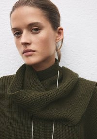 Maglione verde scuro con texture, collo ampio a scialle pieghevole. Presenta costine verticali e una collana con catena tono argento. Sfondo neutro.