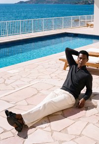 Homme portant un pull noir et un pantalon blanc, allongé sur une terrasse en pierre près de la piscine, avec l'océan et des collines en arrière-plan.