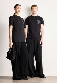 Dwie modelki noszą czarne, krótkie koszulki z nadrukiem, zestawione z luźnymi czarnymi spodniami. Jedna z nich trzyma małą czarną torebkę.