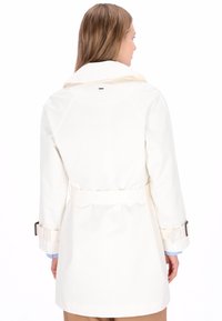 Lysebeige trenchcoat med bælte i taljen og manchet detaljer. Har en bred krave og glat, tekstureret stof. Ren, minimalistisk design.