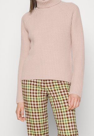 Femme portant un pull à col roulé côtelé rose clair et un pantalon à carreaux taille haute dans des tons marron, beige et vert.